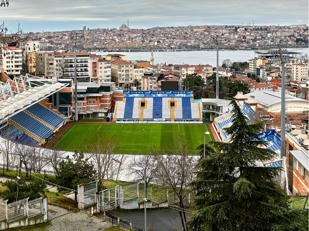 Blick von oben auf ein Fußballstadion mit blau-weißen Tribünen, eingebettet in die Hügel Istanbuls mit dem Bosporus und Moscheen im Hintergrund.